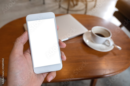 Mockup image of hand holding white mobile phone with blank desktop screen with coffee cup and laptop on table