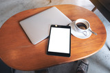 Mockup image of a black tablet pc with blank desktop white screen with laptop and coffee cup on wooden table