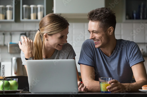 Young happy positive family couple talking laughing during breakfast with laptop, funny cheerful millennial man and woman having fun joking enjoying online computer humor at home in the morning