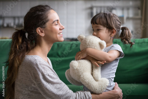 Smiling mother hugging cute little girl holding teddy bear toy showing love and care in family, young mom embracing protecting child, sincere relationships between mum and daughter cuddling concept