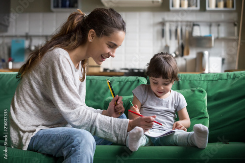 Smiling baby sitter and preschool kid girl drawing with colored pencils sitting on sofa together, single mother and child daughter playing having fun, creative family activities at home concept photo