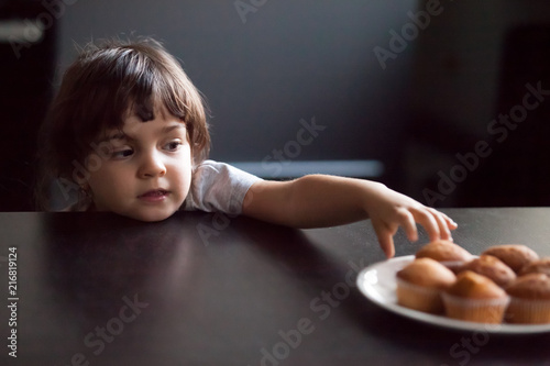 Obraz na plátně Cunning cute little girl stealing delicious muffin on table, hungry funny impati