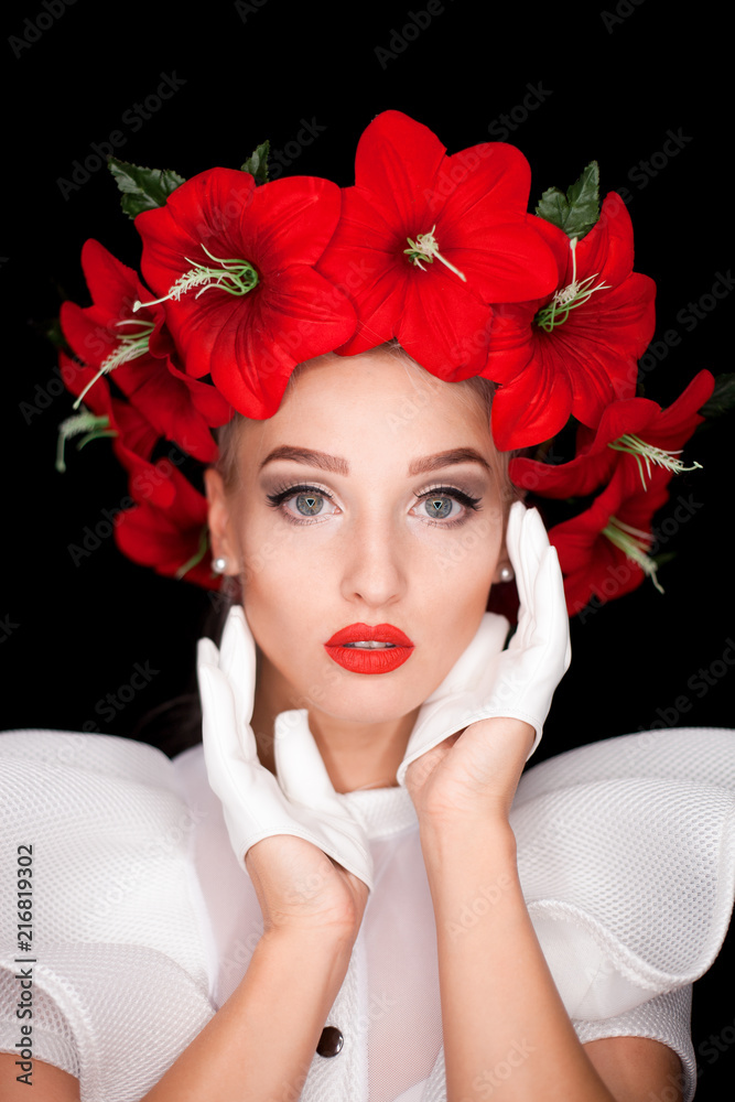 Naklejka premium Portrait of a girl in white with a wreath of red flowers on her head, holding her hands in white gloves next to her face on a black background