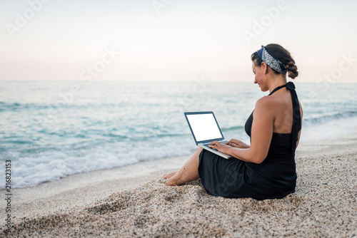 Freelance work concept. Young woman using laptop on the beach near the water. © bnenin
