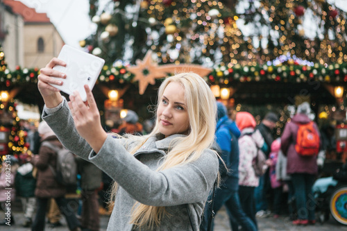Photography A beautiful young blonde woman or girl doing selfie or photographing next to a Christmas tree during Christmas holidays at the Old Town Square in Prague, Czech Republic