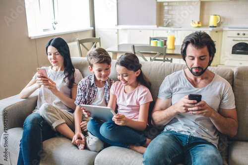 Parents are sitting on sofa with kids and look at phones. Children are in bettween woman and man. Girl holds tablet in hands. They look at screen.