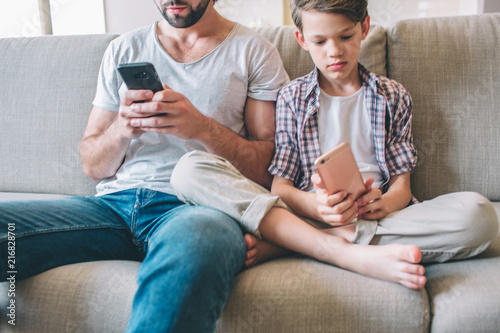 Cut view of man and boy sitting on sofa and looking at phone they have in hands. Child sits with legs corssed. photo