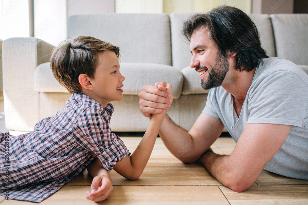 A positive and nice picture of father and son lying on the floor and holding each other's hands. They are playing. A positive and nice picture of father and son lying on the floor and holding each other's hands. They are playing.