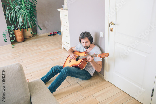 A picture of gy sitting alone on the floor and playing the guitar. He is leaning to the wall. Man is entertaining himself. photo