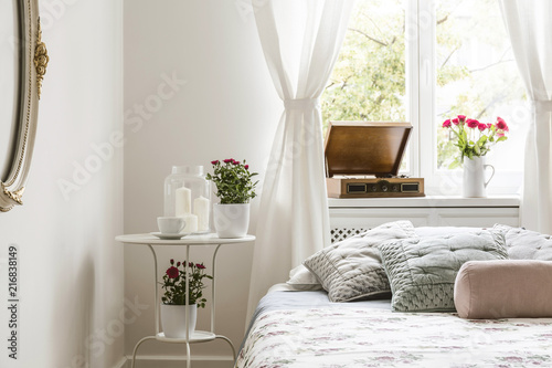 White bedroom interior in real photo with bedside table with tea cup, candles and red roses, windowsill with gramophone and fresh flowers