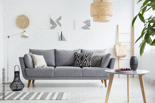Real photo of a scandi living room interior with cushions on gray couch, cherries on wooden table and bag on a ladder photo