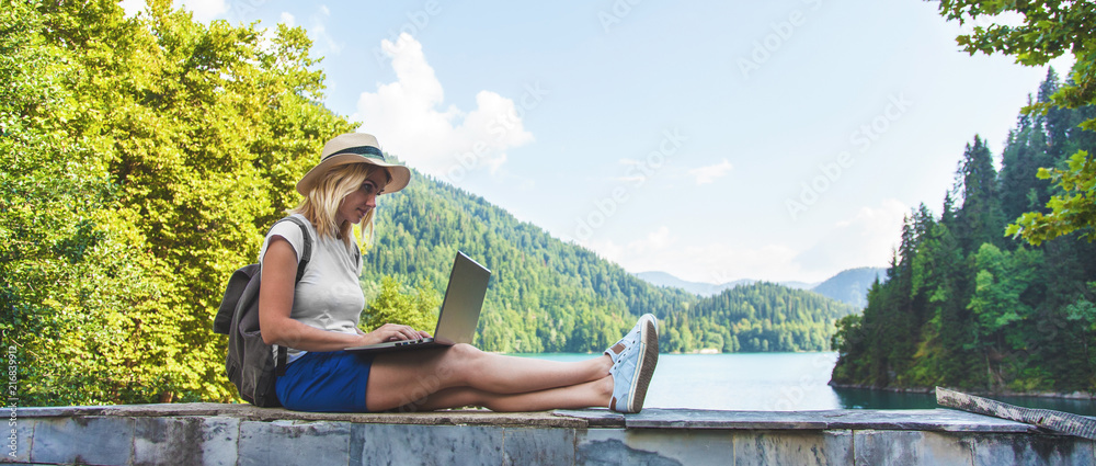 Beautiful girl traveler in a hat on a lake with a laptop Work outside the office - obrazy, fototapety, plakaty 