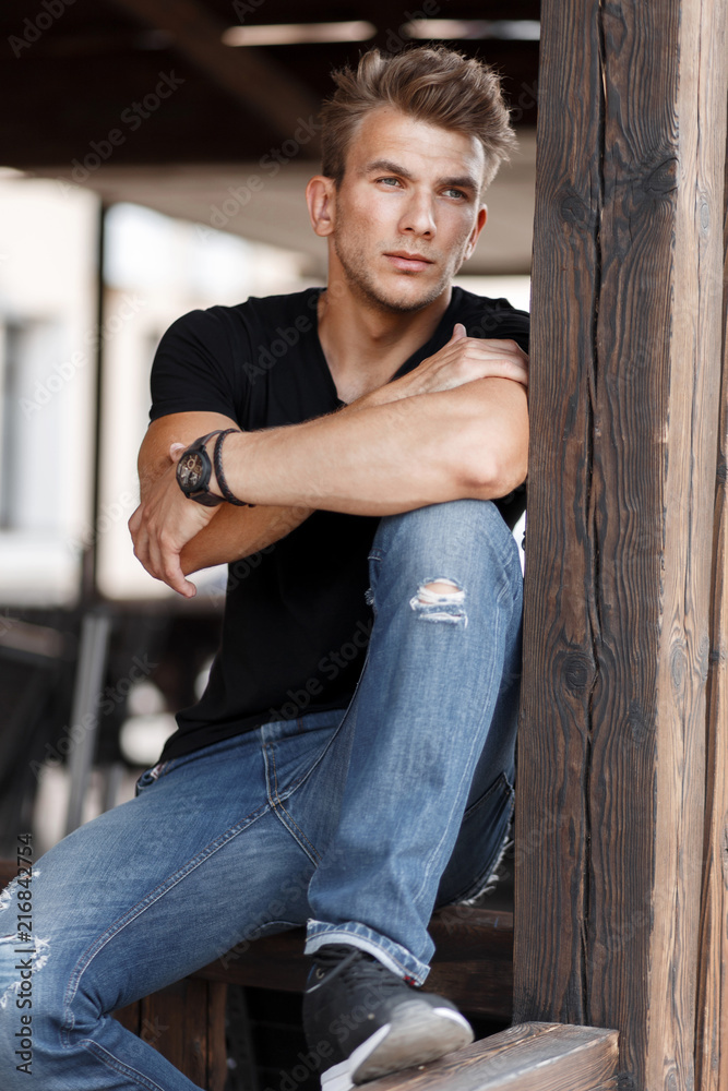 handsome young man with a hairstyle in a fashionable black T-shirt and blue  jeans sits on a wooden building Stock Photo | Adobe Stock