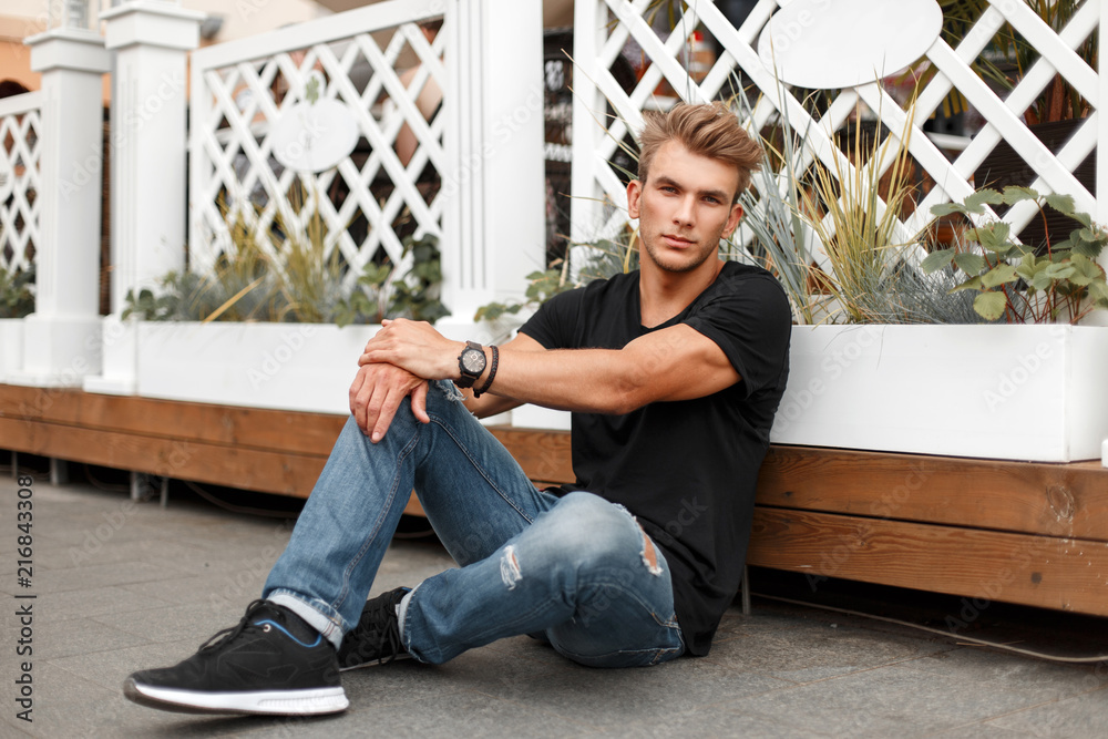 Fashionable handsome young man model with a hairstyle in a black T-shirt and jeans with sneakers sits near a wooden fence - obrazy, fototapety, plakaty 