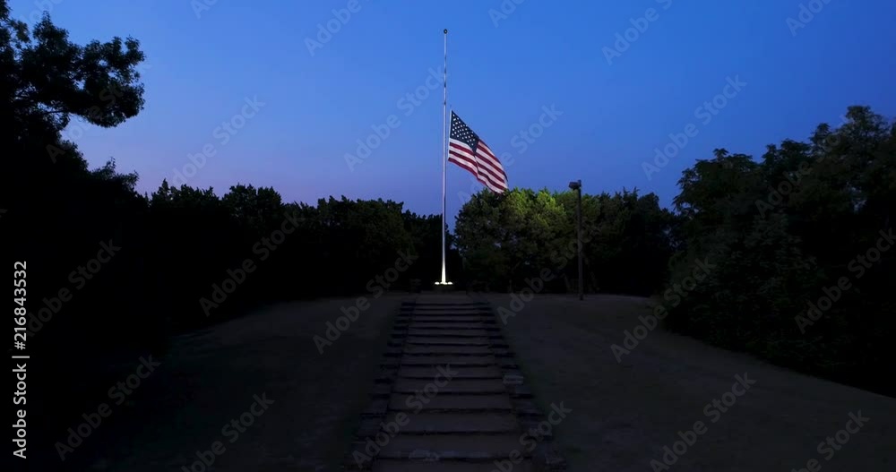 Drone view of US flag at half-staff at dawn, lit by spotlight,, moving forward and up