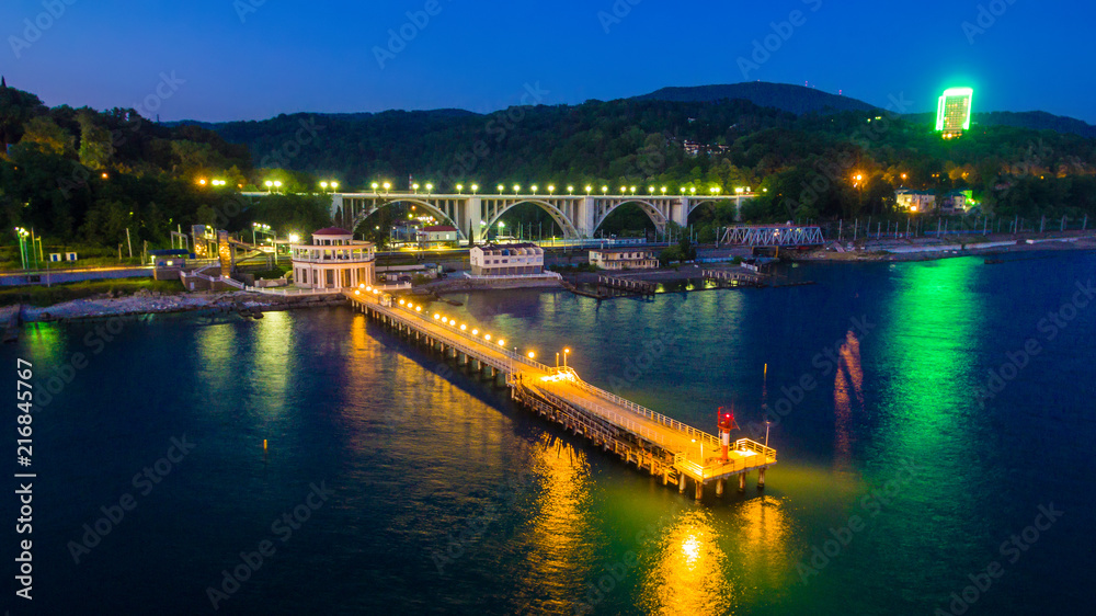 Drone view of the pier of the Matsesta marine station on the background of the illuminated Matsesta viaduct and mountains at twilight, Sochi, Russia
