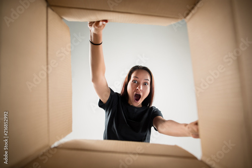 The surprised woman unpacking, opening carton box and looking inside. The package, delivery, surprise, gift, lifestyle concept. Human emotions and facial expressions concepts