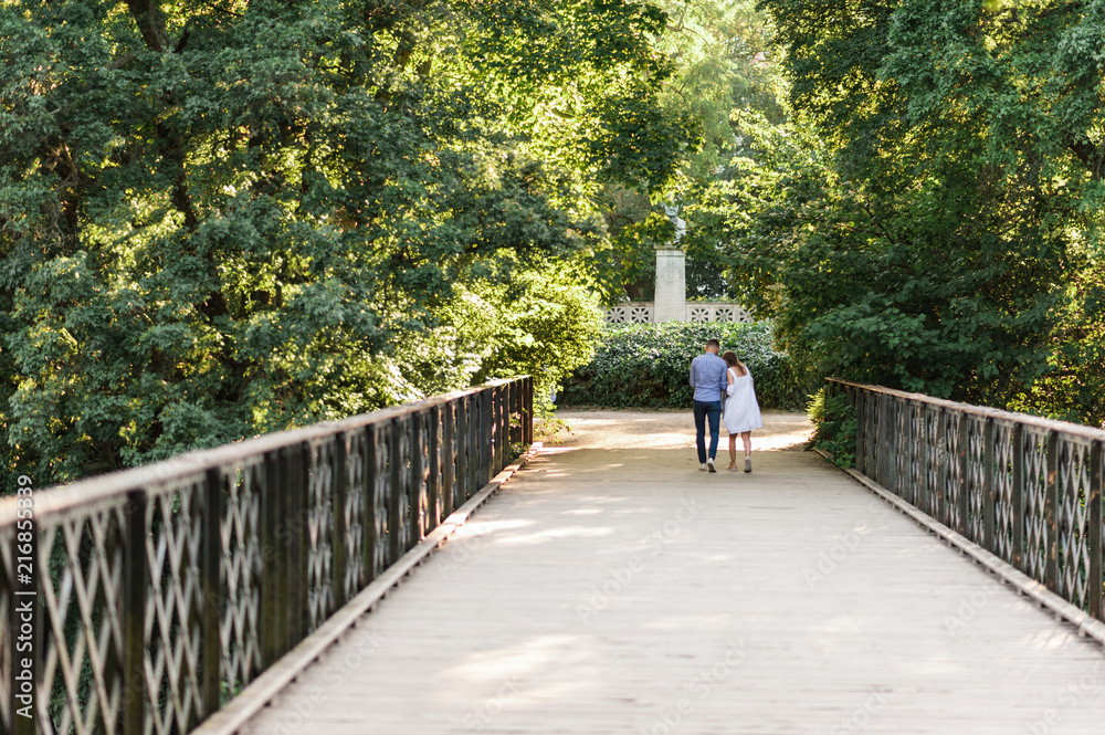 Young couple walking along peaceful park scenery on a beautiful summer day. Green canopy of trees and a metal bridge above the water of a pond. Concept of hygge.