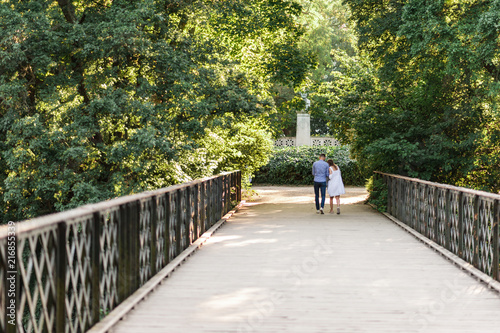 Young couple walking along peaceful park scenery on a beautiful summer day. Green canopy of trees and a metal bridge above the water of a pond. Concept of hygge.