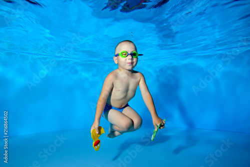A happy little boy swims and poses underwater in the pool on a blue background with swimming glasses and toys in his hands. He looks at the camera and smiles. Portrait. Underwater photography