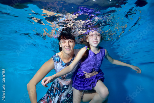 Happy mom and daughter swim and pose underwater in the pool in beautiful dresses. They look at the camera and smile. Portrait. Underwater photography. Horizontal orientation