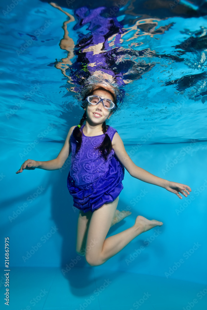Happy little girl with pigtails swims and poses underwater in the pool in swimming glasses and purple dress. She looks at the camera and laughs. Portrait. Shooting underwater. Vertical view