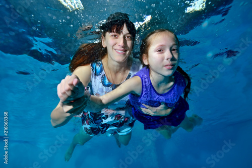 Mom and daughter swim underwater in the pool in beautiful dresses. Mom helps daughter. They look at the camera and smile. Portrait. Close up. Underwater photography