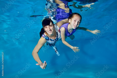 A little girl in a purple dress with her mother swims underwater in the pool on a blue background. They hug, look at the camera and smile. Portrait. Underwater photography