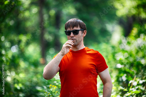 A young white bearded man in red t-shirt and in sunglasses is vaping an electronic cigarette in the forest in the summer. Close up.