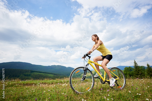 Athlete woman cyclist riding on yellow mountain bicycle on a grassy hill, enjoying summer day in the mountains. Outdoor sport activity, lifestyle concept