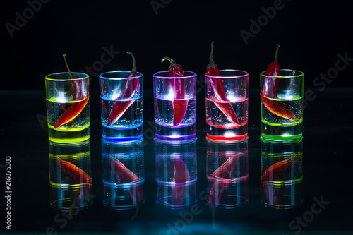 Five multicolored shot glasses full of drink and with the red chili peppers lying inside them symmetrically placed a black background.
