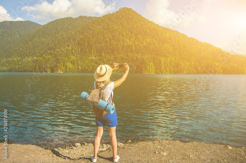 Beautiful girl traveler in a hat stands on a lake and takes pictures on a background of mountains Travel concept Summer vacation Copy space