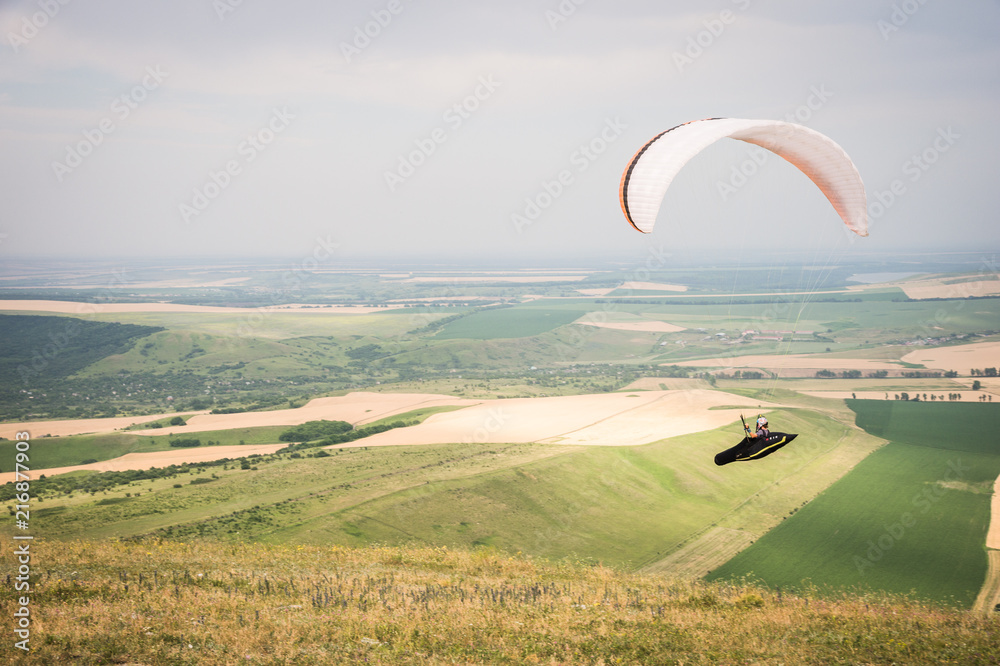 White orange paraglide with a paraglider in a cocoon against the background of fields of the sky and clouds. Paragliding Sports White orange paraglide with a paraglider in a cocoon against the background of fields of the sky and clouds. Paragliding Sports