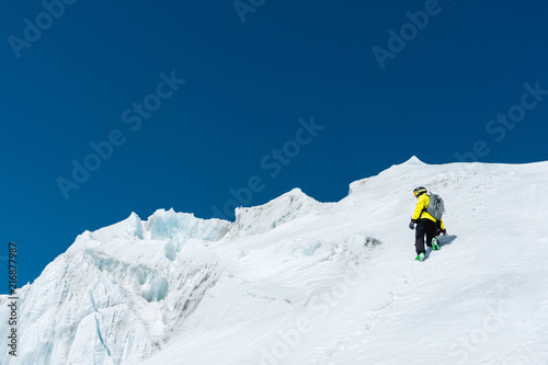 A skier in a helmet and mask with a backpack rises on a slope against the background of snow and a glacier. Backcountry Freeride