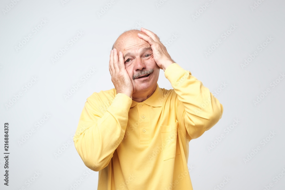 Senior hispanic man in yellow tshirt pulling skin on face with hands. He is scared or tired of stress. Senior hispanic man in yellow tshirt pulling skin on face with hands. He is scared or tired of stress.