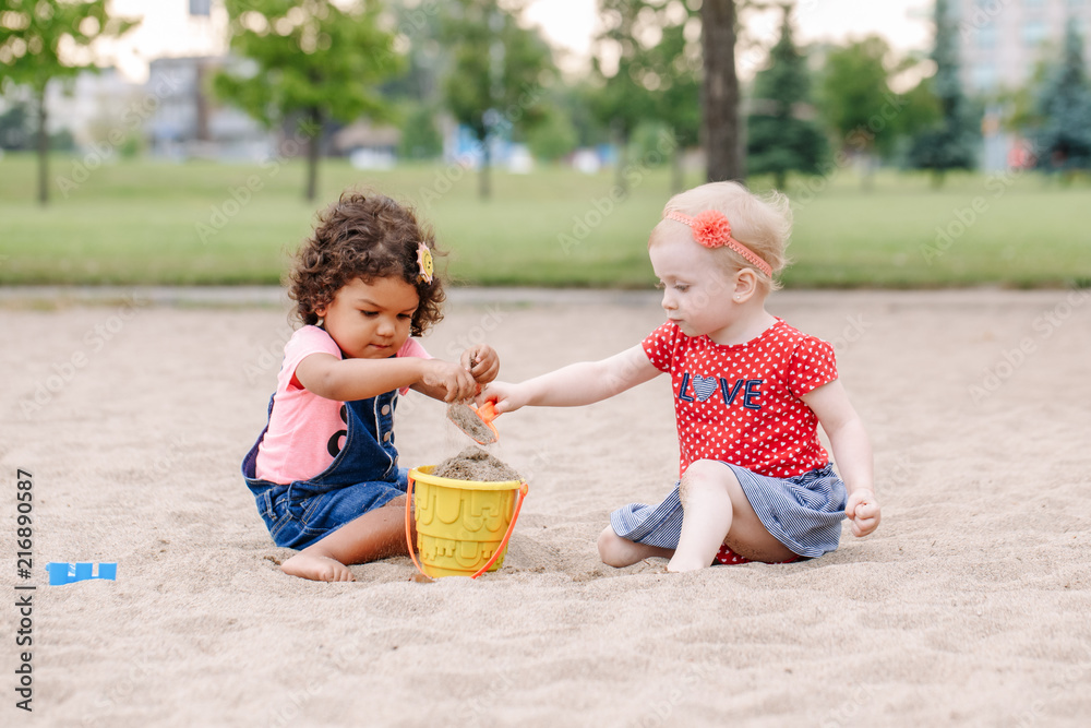Portrait of two cute Caucasian and hispanic latin toddlers babies children sitting in sandbox playing with plastic colorful toys. Little girls friends having fun together on playground. Portrait of two cute Caucasian and hispanic latin toddlers babies children sitting in sandbox playing with plastic colorful toys. Little girls friends having fun together on playground.