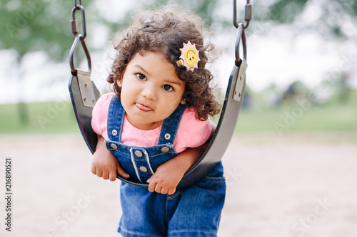 Portrait of happy smiling little latin hispanic toddler girl swinging on swings at playground outside on summer day. Happy childhood lifestyle concept. Toned with film pastel faded filters colors.