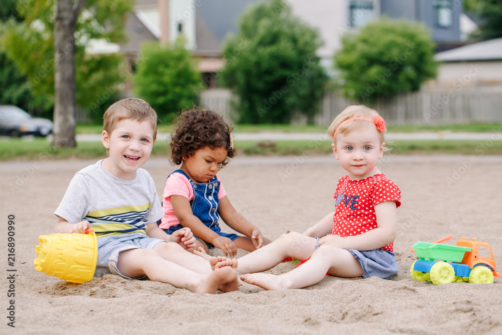 Portrait of three cute Caucasian and hispanic latin toddlers babies children sitting in sandbox playing with plastic colorful toys. Little girls and boy friends having fun together on playground. Portrait of three cute Caucasian and hispanic latin toddlers babies children sitting in sandbox playing with plastic colorful toys. Little girls and boy friends having fun together on playground.