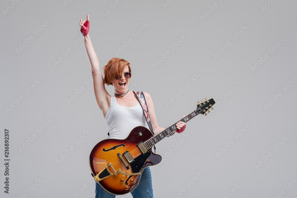 Fototapeta premium Portrait of a young woman musician with an electric guitar in her hands on a gray background. She wears black sunglasses, laughs and plays rock and roll loudly