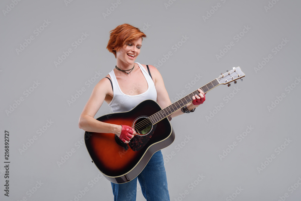 Young woman musician with an acoustic guitar in hand on a gray background. He laughs and plays rock and roll loudly. Young woman musician with an acoustic guitar in hand on a gray background. He laughs and plays rock and roll loudly.