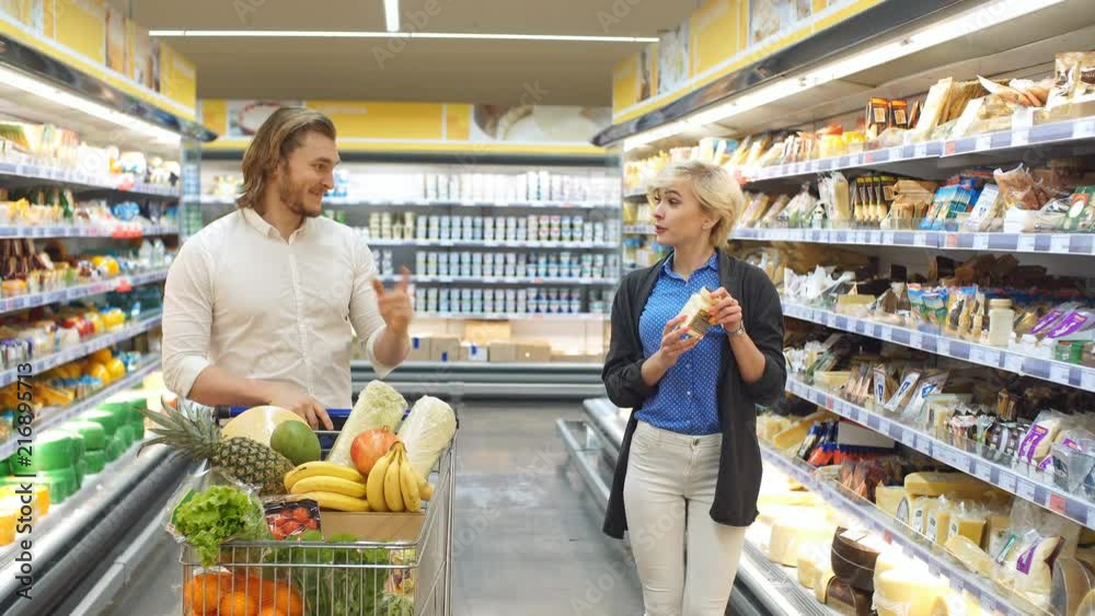 Caucasian cheerful male and female shoppers push cart with grocery products in supermarket aisle