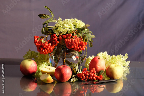 Still life with apples and ashberries on the background of a jug of flowers is reflected on the glass.