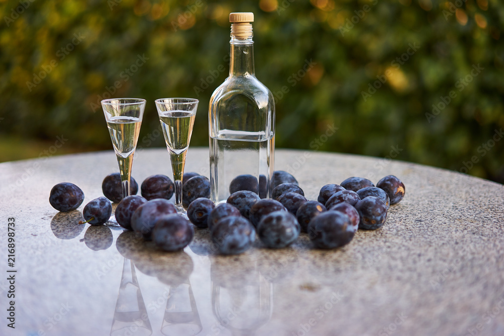 Stockfoto Still life Picture of the bottle of homemade slivovitz or plum  brandy with two glases of short drink and fresh riped plums on the stone  table in garden restaurant. | Adobe