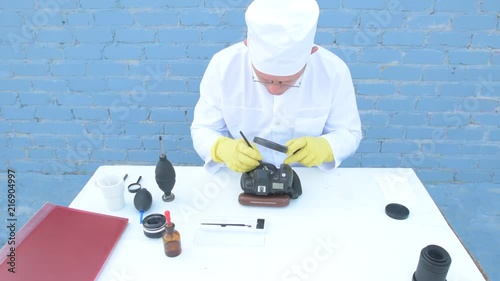 Elderly solid person in white dressing gown, cap, medical mask, glasses cleans matrix of digital camera with special means. Lens, sensor, matrix, cleaning wands, magnifier, isopropyl alcohol. photo