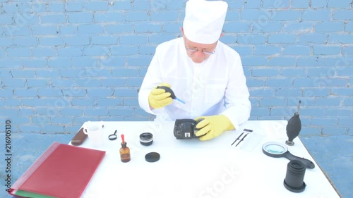 Elderly solid person in white dressing gown, cap, medical mask, glasses cleans matrix of digital camera with special means. Lens, sensor, matrix, cleaning wands, magnifier, isopropyl alcohol. photo