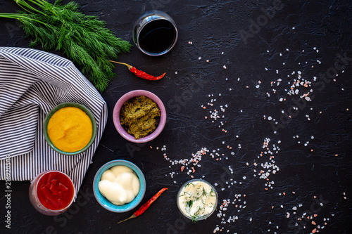 Spicy sauses. Ketchup, mayonnaise, mustard, soy sauce, barbecue sauce, pesto, mustard, sour near chili pepper and greenery on black background top view space for text