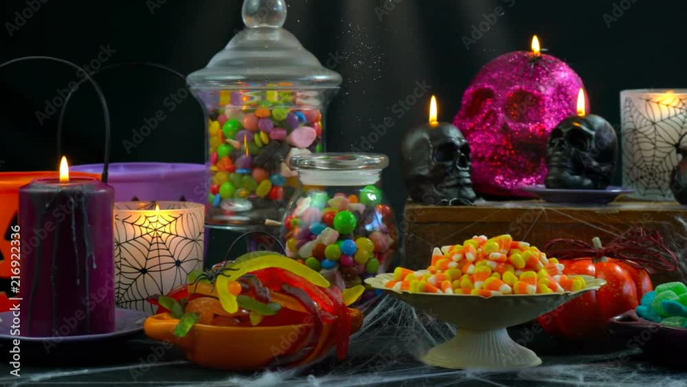 Happy Halloween trick or treat party table with bowls and apothecary jars of candy with skull candles against a black background, scooping candy close up. Happy Halloween trick or treat party table with bowls and apothecary jars of candy with skull candles against a black background, scooping candy close up.