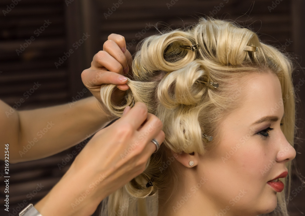 Fototapeta premium elegant blonde woman in beauty saloon. Hairdresser does coiffure in the form of big curl. Concept wedding hairstyle