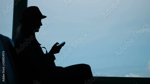 Cinemagraph of Female Traveler Texting on Mobile Phone While Waiting in Lounge with Moving Clouds at Backgroud photo