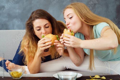 compulsive overeating  mindless snacking  sedentary lifestyle  hunger  pleasure  enjoyment. Two young women sitting on coach greedily engorging burgers and chips